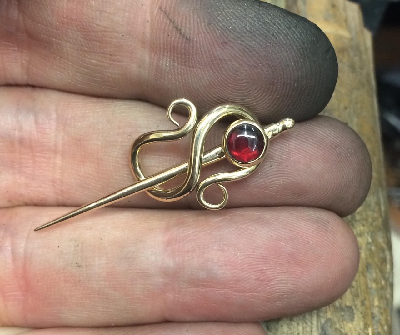 A gold cloak pin with a red stone, held in a silversmith’s work smudged fingers.