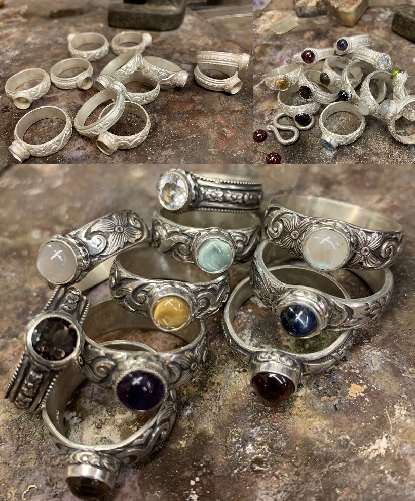 Handcrafted sterling silver rings with gemstones, piled on a dark workbench.